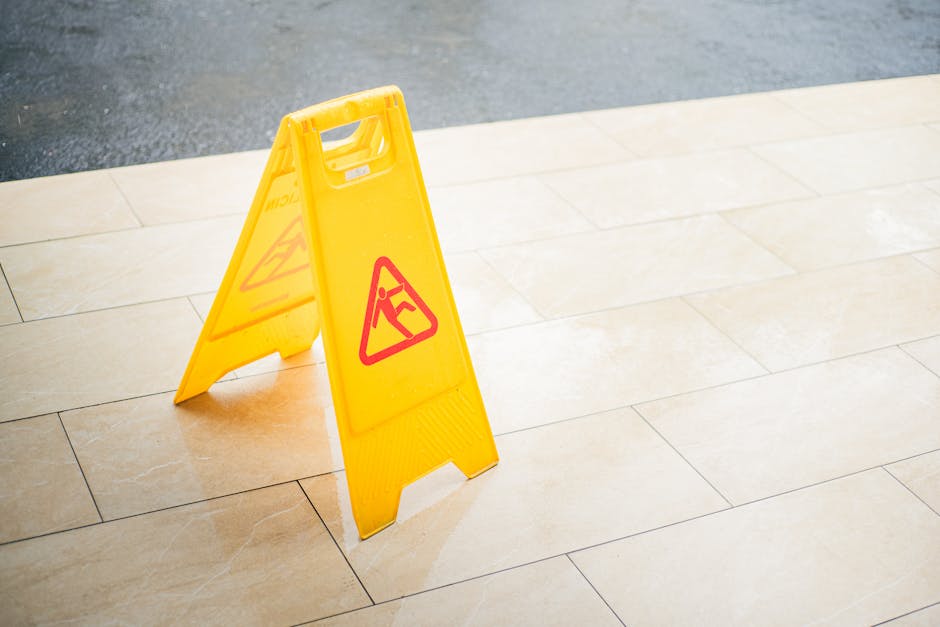 A bright yellow, collapsible warning sign with red triangular symbols indicating a slipping hazard, placed on a beige tiled floor near the edge of the pavement. The sign is standing upright, with a sm